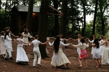 a video still of a group of women dressed in white, holding hands, dancing around a tree. By Colina van Bemmel